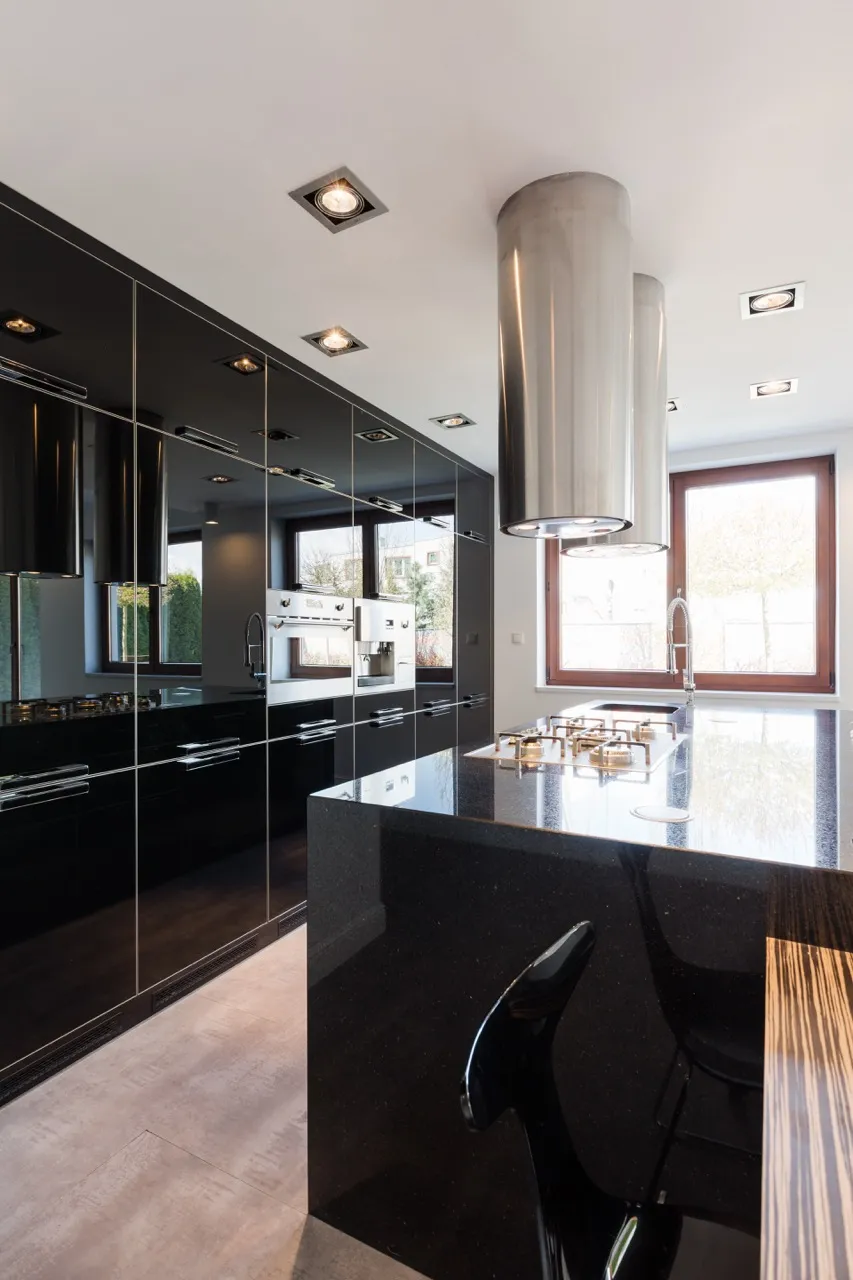 Modern kitchen with glossy black cabinets, built-in stainless steel ovens, a sleek island cooktop, and a large range hood. Perfect for Home Remodeling Boca Raton, this bright space is completed by a stylish chair and abundant natural light.